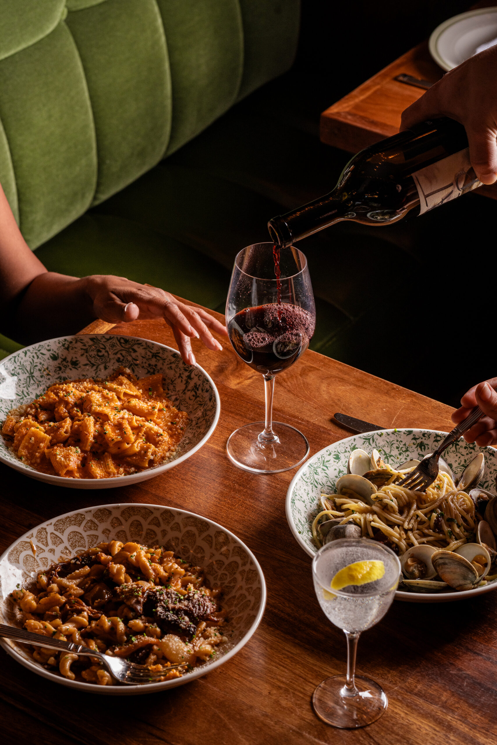 A person's hand holding a glass of red wine above a wooden table with a bowl of pasta and a bowl of seafood.